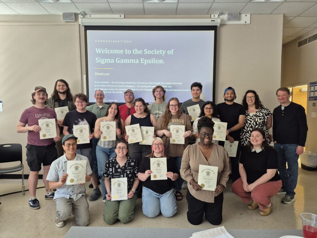 Student and faculty charter members pose in 3 rows in front of a projection slide Welcoming the Iota Eta Chapter