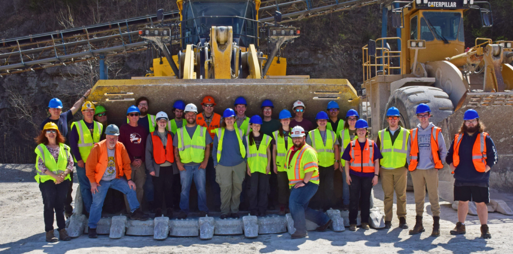 Group members wearing safety clothing pose in the bucket of a loader